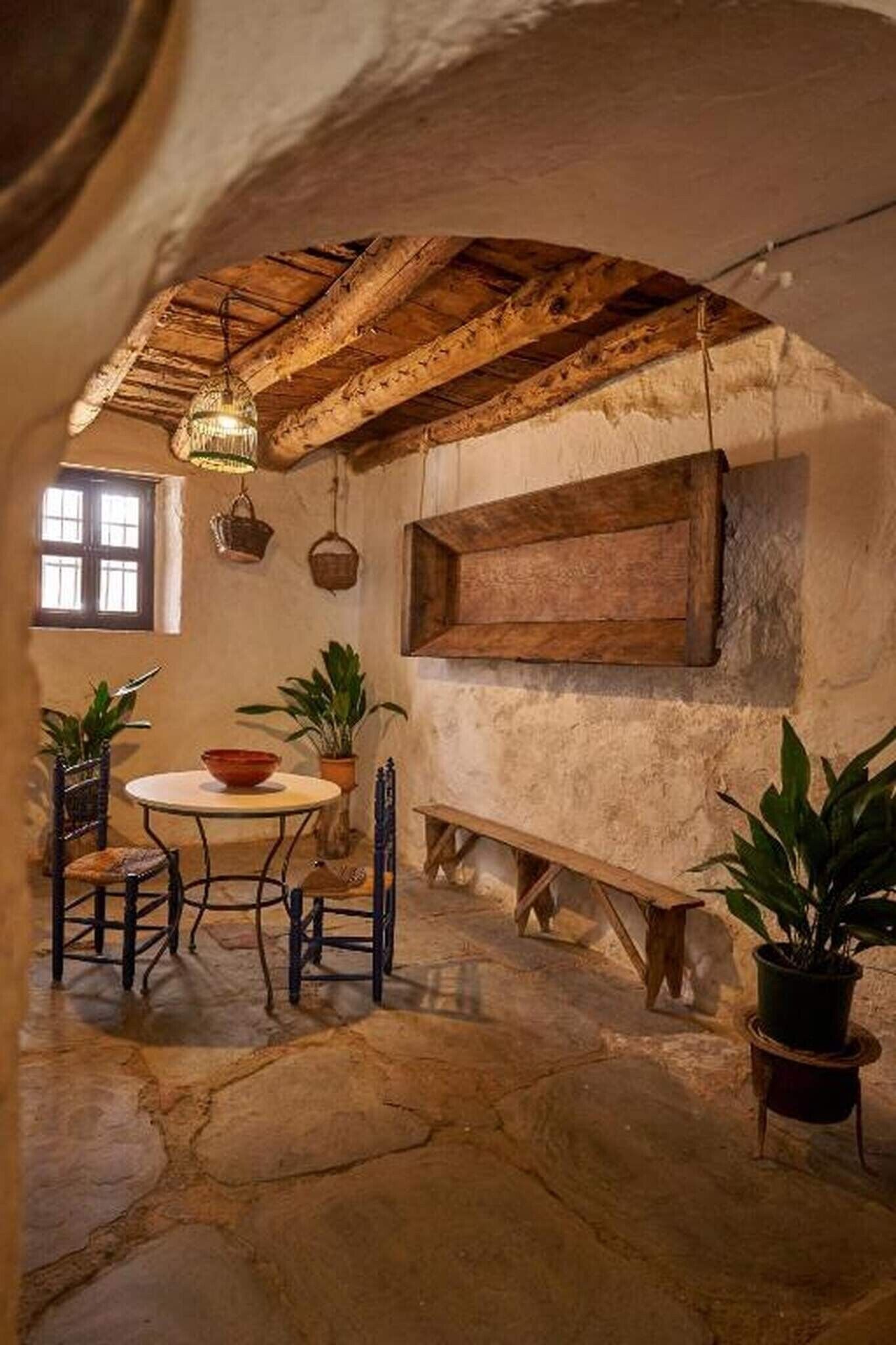 Un intérieur rustique avec un sol en pierre, des poutres en bois, une table ronde entourée de chaises et des plantes en pot.