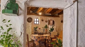 A rustic dining area with a wooden table and chairs, potted plants, and a stone floor.