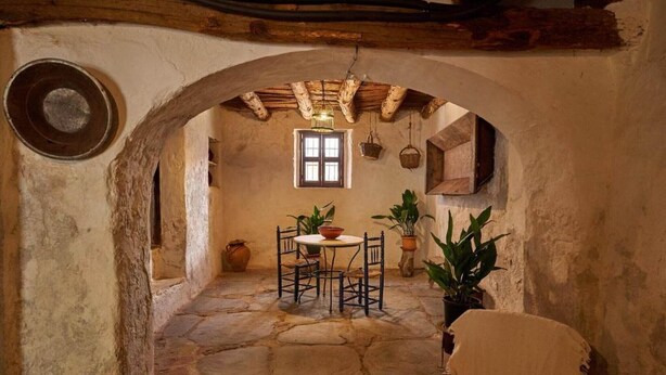An arched stone room with a table and chairs, a hanging light fixture, and potted plants.