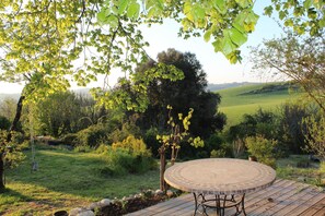 A circular outdoor seating area with a stone table and metal chairs, surrounded by a wooden deck and a grassy landscape with trees.