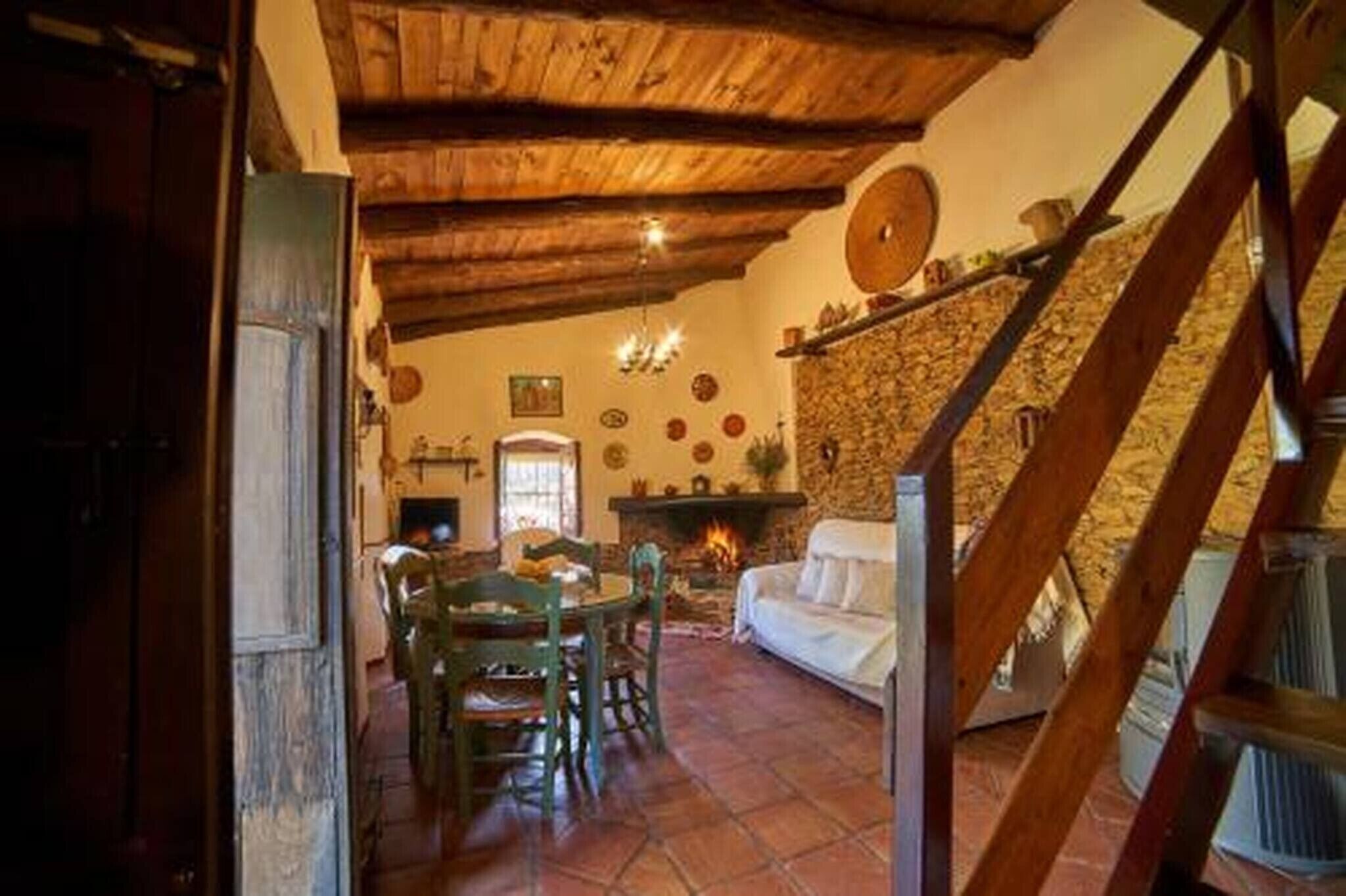 Un interior rústico con pared de piedra, techo de madera, chimenea, zona de comedor y una escalera.
