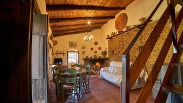Un interior rústico con pared de piedra, techo de madera, chimenea, zona de comedor y una escalera.