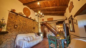A cozy room with a stone wall, wooden ceiling, a sofa, a dining table with chairs, and a staircase leading to another area.