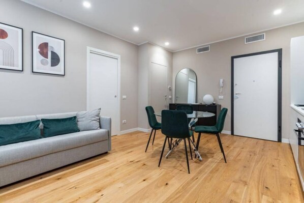 Un salon moderne avec un canapé gris, des coussins verts, une table à manger avec des chaises et un grand miroir au mur.