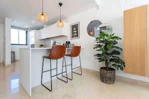 Una cocina moderna con armarios blancos, una isla central con taburetes, iluminación colgante, un espejo grande y una planta en maceta.