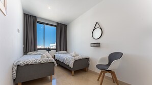 Ein modernes Schlafzimmer mit zwei Betten, einem Sessel, einem großen Fenster und einem Wandspiegel.