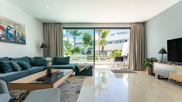 Ein modernes Wohnzimmer mit Sofa, Couchtisch, Fernseher und einem Balkon mit Gartenmöbeln.
