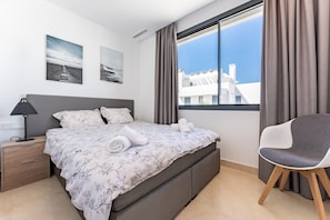 Un dormitorio con cama, una silla, una mesita de noche y una ventana con cortinas.
