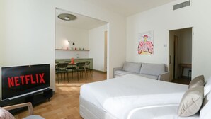 Ein Hotelzimmer mit einem großen Bett, einem Fernseher der 'NETFLIX' anzeigt, einer Küchenzeile und einem Essbereich.