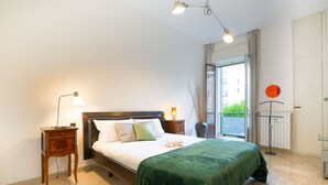 A bedroom with a bed, bedside lamps, a radiator, and a view of greenery outside.