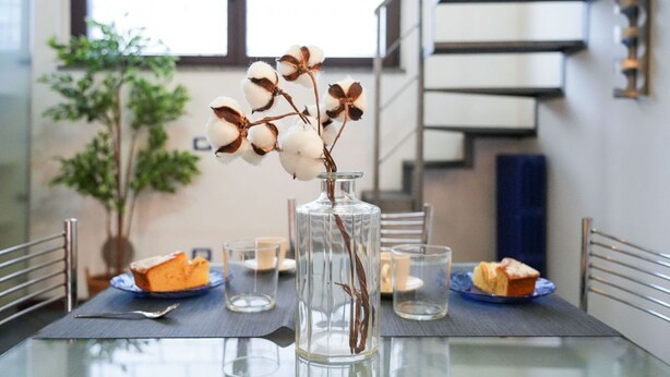A dining table with a clear glass vase containing cotton branches, surrounded by plates with food and candles. - Milano, Industrial Loft (Milano)