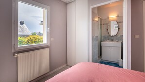 Et soveværelse med en lyserød seng, et vindue med udsigt til bygninger og blomster, en radiator, samt et badeværelse med spejl og vask.