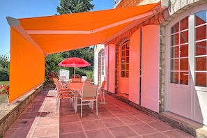 Une terrasse avec une table et des chaises, un parasol rouge et une maison à la façade en pierre.