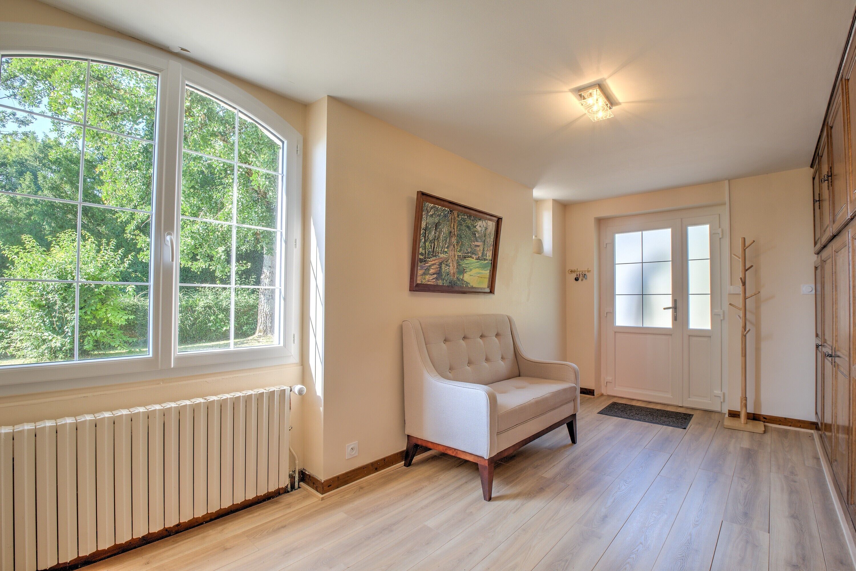 Una habitación con un amplio ventanal, un radiador blanco, un sofá color beige, una puerta con paneles de vidrio esmerilado y un cuadro en la pared.