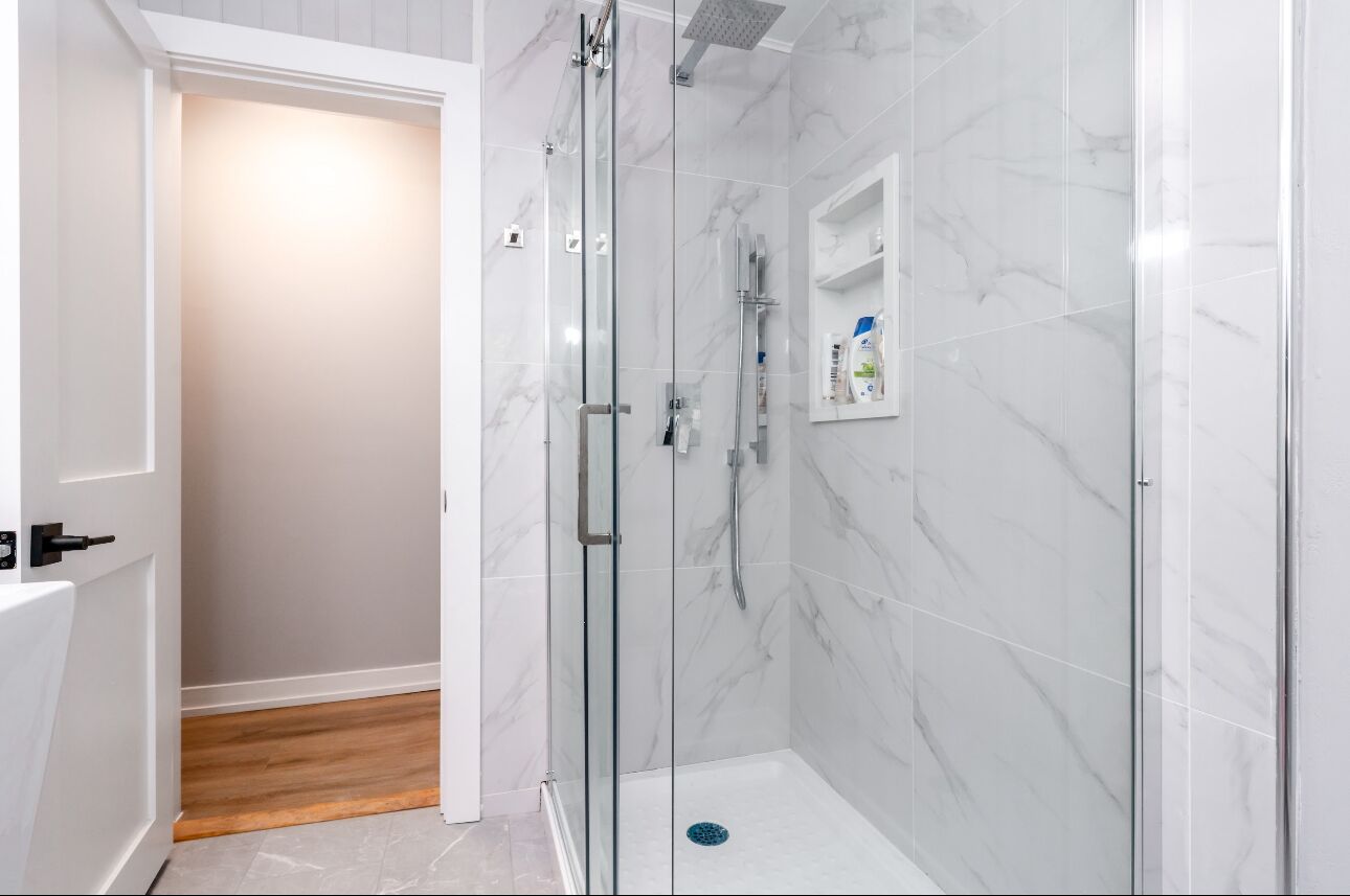 A modern bathroom with a glass-enclosed shower, marble walls, and a wooden floor.