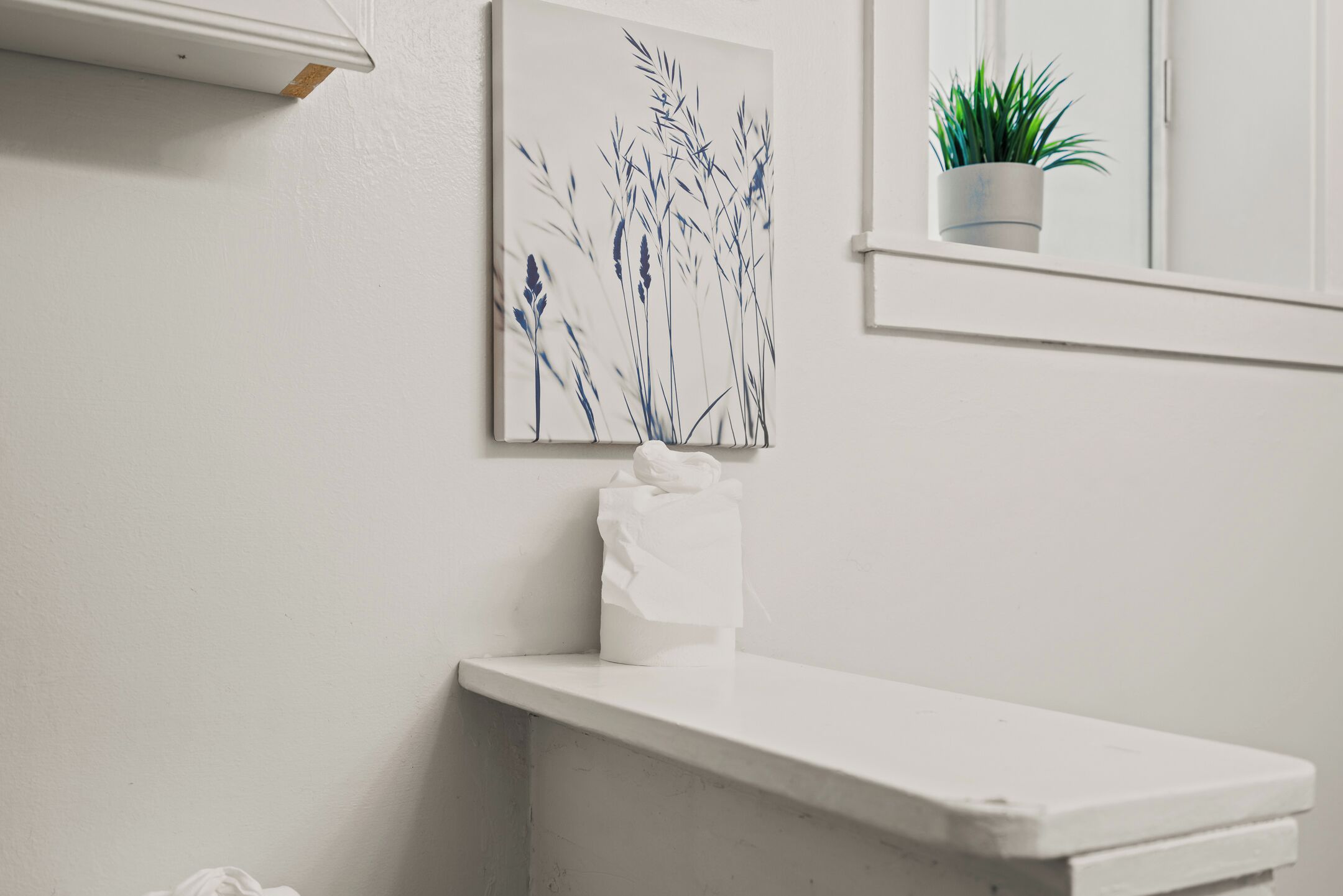 Une salle de bain avec un lavabo blanc, un distributeur de serviettes blanc, une œuvre d’art encadrée représentant des herbes hautes et une plante en pot posée sur le rebord de la fenêtre.