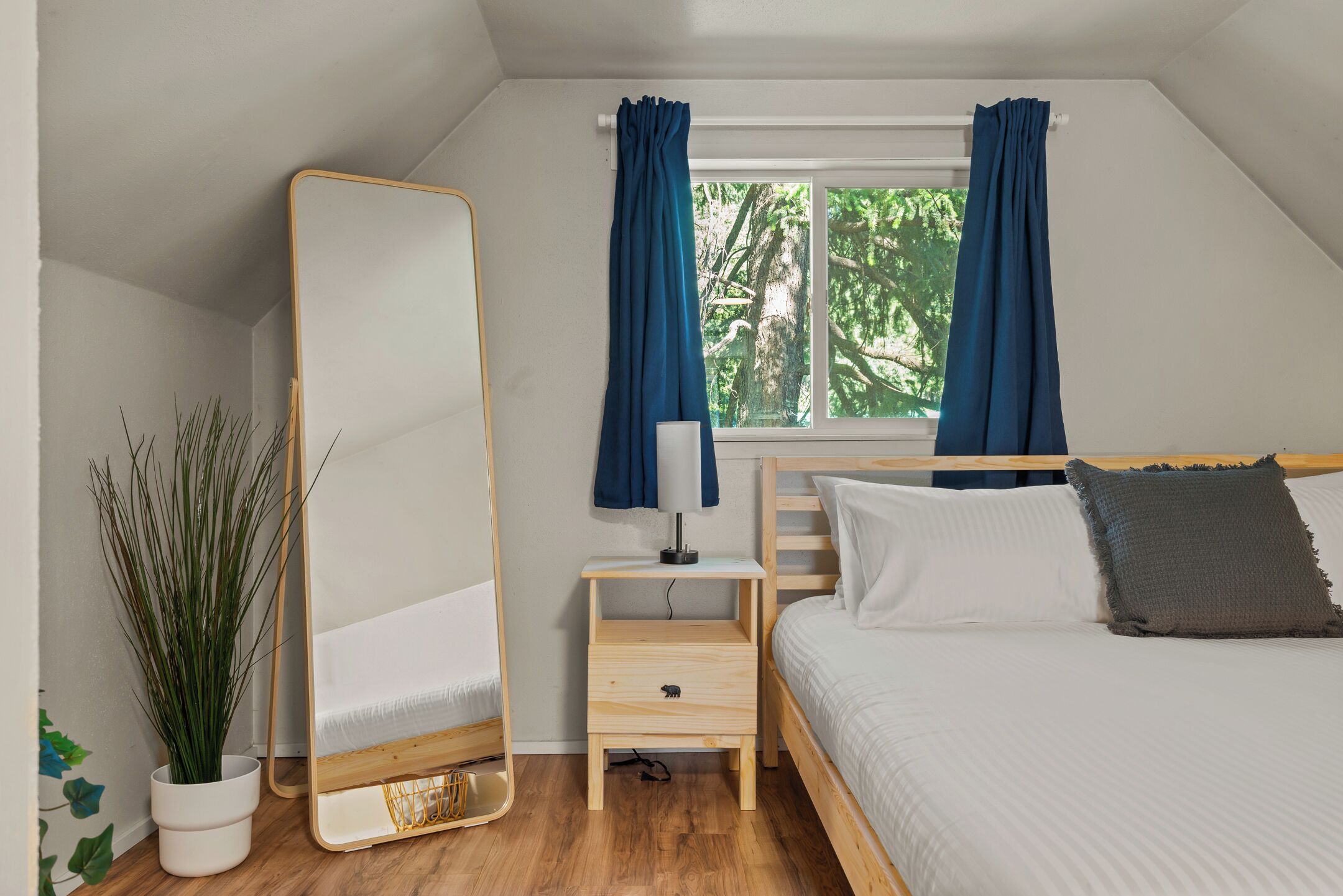 A bedroom with a bed, a bedside table with a lamp, a mirror, and a window with curtains.