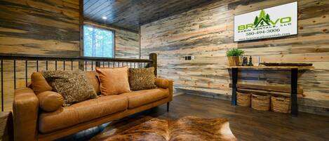Interior de una cabaña con techo de madera, un sofá de cuero con cojines, un manto de piel y una pared de madera con un logotipo e información de contacto.