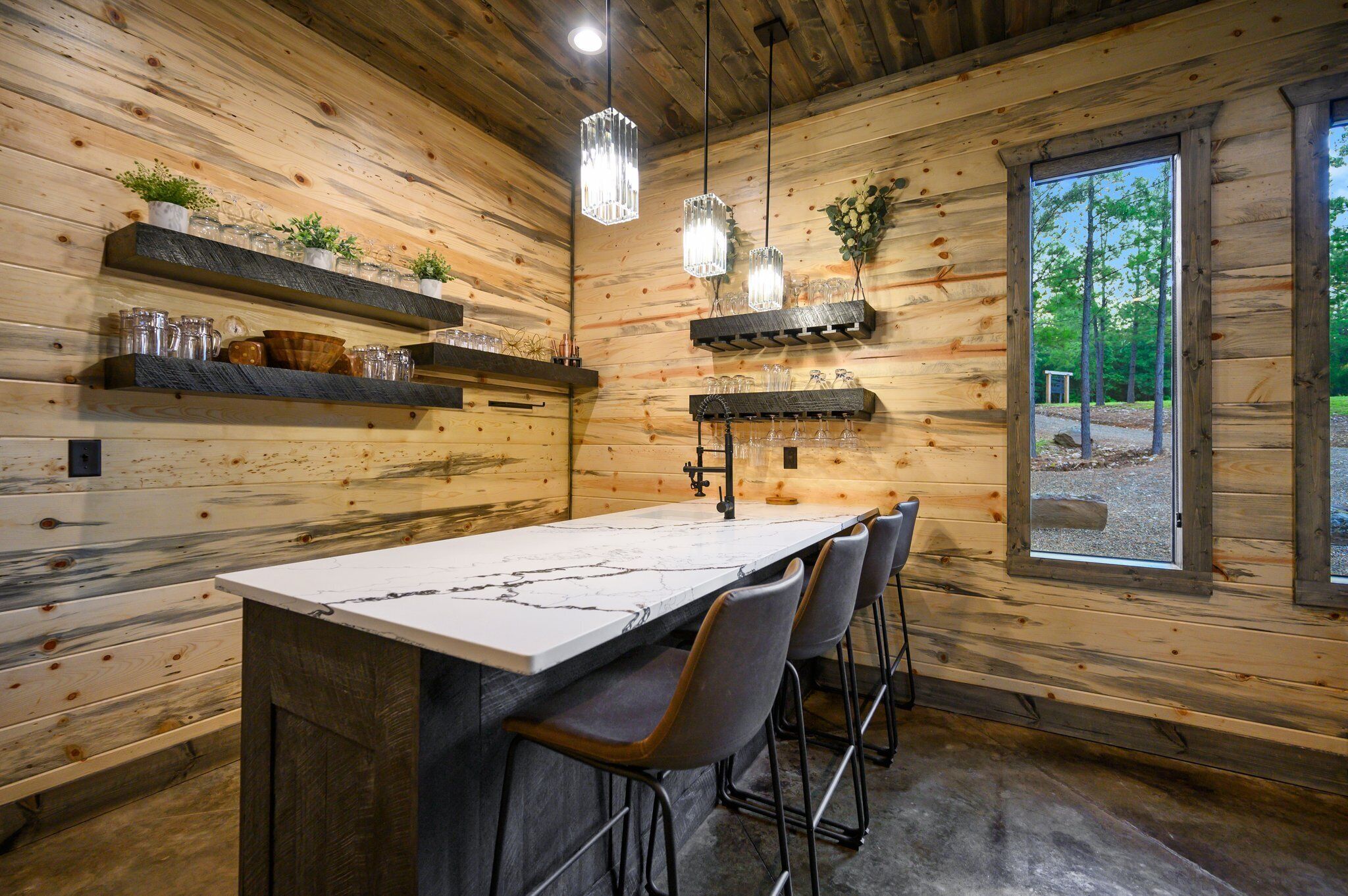 A wooden cabin interior with a marble bar, shelves with glassware, and a view of the outdoors.