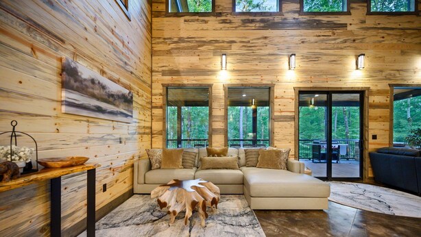 A modern log cabin interior with a sectional sofa, a wooden coffee table, and large windows offering a view of the forest. - Runaway Streams - Game Room, Mini Golf, Creekside, EV Outlet (Broken Bow)