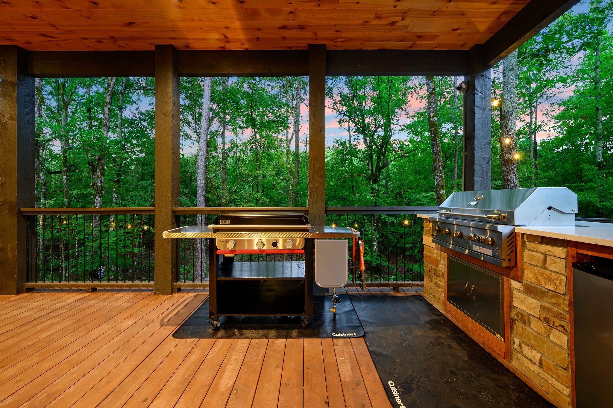 An outdoor kitchen with a grill and oven, surrounded by a wooden deck and a railing, overlooking a forested area.