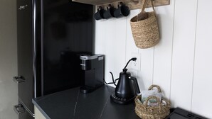 Una cocina con un refrigerador negro, una cafetera y un hervidor sobre el mostrador. Hay un estante de madera encima del mostrador con una cesta colgante y una lámpara.