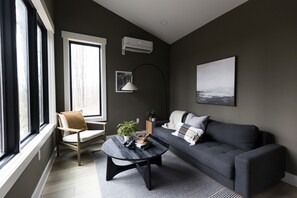 Una sala de estar moderna con un sofá gris, una mesa de centro redonda y un ventanal que proporciona luz natural.