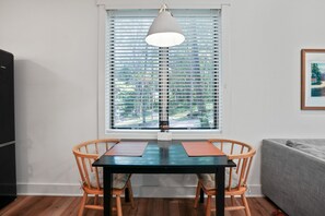 Un coin repas avec une table et des chaises, une fenêtre avec des stores, un luminaire suspendu et un tableau encadré au mur.