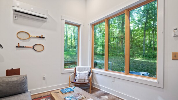 Una habitación con ventanas amplias, una mesa de madera con libros, una silla y raquetas de tenis en la pared.