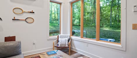 A room with large windows, a wooden table with books, a chair, and tennis rackets on the wall.