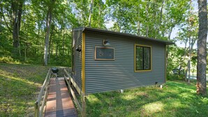 A small cabin with a wooden deck leading to a grill area, surrounded by a forest.