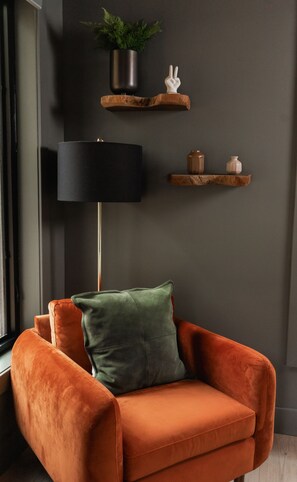 En moderne stue med væg i mørkegrå, gulvlampe i sort, træhylde med plante, lille hvid figur og grøn pude på en orange velourstol.