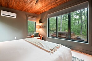 Une chambre avec un grand lit, un plafond en bois, une fenêtre donnant sur la forêt et un petit coin salon avec une lampe.