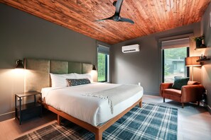 Un dormitorio con techo de madera, cama con ropa de cama blanca, alfombra a cuadros, una mesita de noche, una silla y una ventana con vistas a zonas verdes.