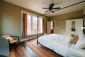 A bedroom with a four-poster bed, a ceiling fan, a window with curtains, a chair, and a door leading to another room. - Ivy Cottage | Hocking Hills, Modern, Hot Tub (Creola)