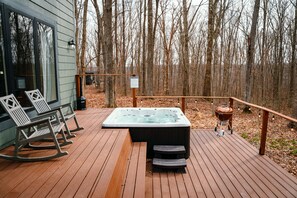 Une terrasse en bois avec un jacuzzi, un barbecue, des fauteuils à bascule et une porte vitrée donnant sur une pièce.