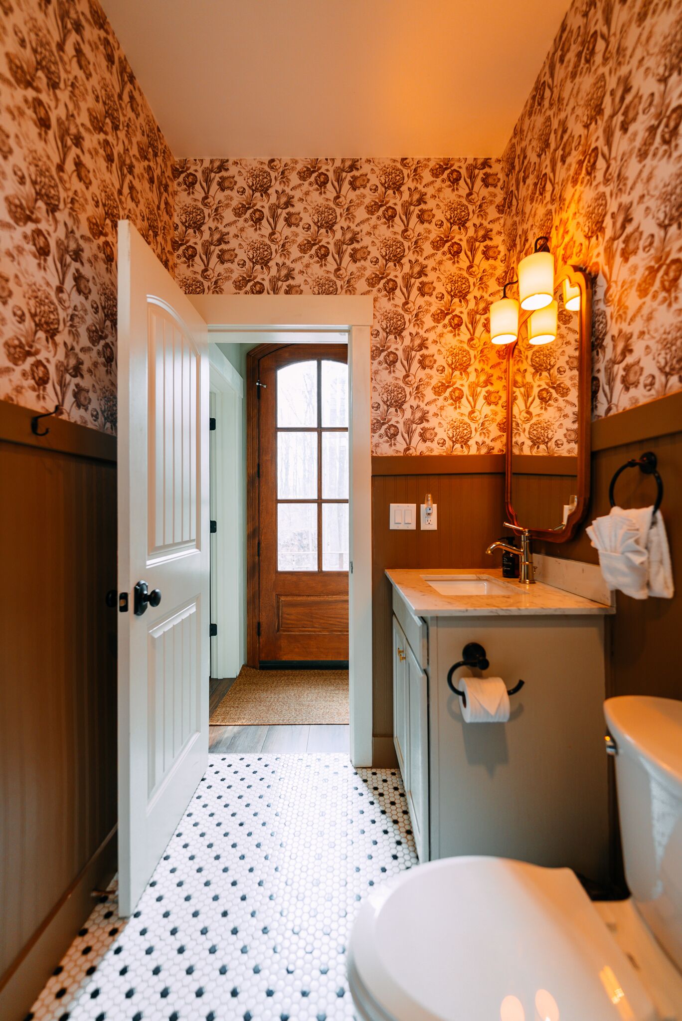 Une salle de bain avec un papier peint à motifs, une porte blanche, un lavabo avec un miroir et des toilettes.