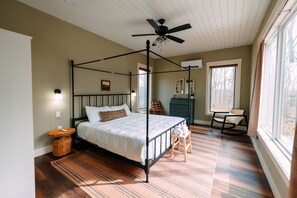 A four-poster bed with a ceiling fan, a wooden floor, a small side table, and a chair by a window.