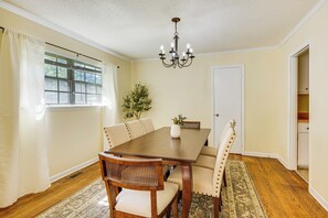 Un comedor con una mesa de madera, sillas tapizadas en tono beige, una araña y una ventana con cortinas.