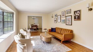 Un salon avec un canapé beige, un fauteuil blanc, une table basse ronde et un tapis à motifs. On peut voir des tableaux encadrés au mur, un lustre et, au fond, un coin repas.