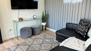 A modern living room with a flat-screen TV, a glass coffee table, two grey ottomans, a patterned rug, a black leather sofa with decorative pillows, and a neon sign on the wall.