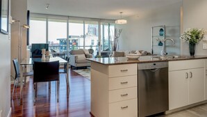 Une cuisine moderne équipée d’un lave-vaisselle, un coin repas avec une table et des chaises en verre, et une vue sur la ville grâce à de grandes fenêtres.