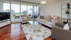 Un salon moderne comprenant un canapé, des fauteuils, une table basse et un téléviseur. De grandes fenêtres offrent une vue sur la ville.