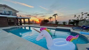 Piscina com boias de unicórnio infláveis, um alpendre coberto e um pôr do sol ao fundo.