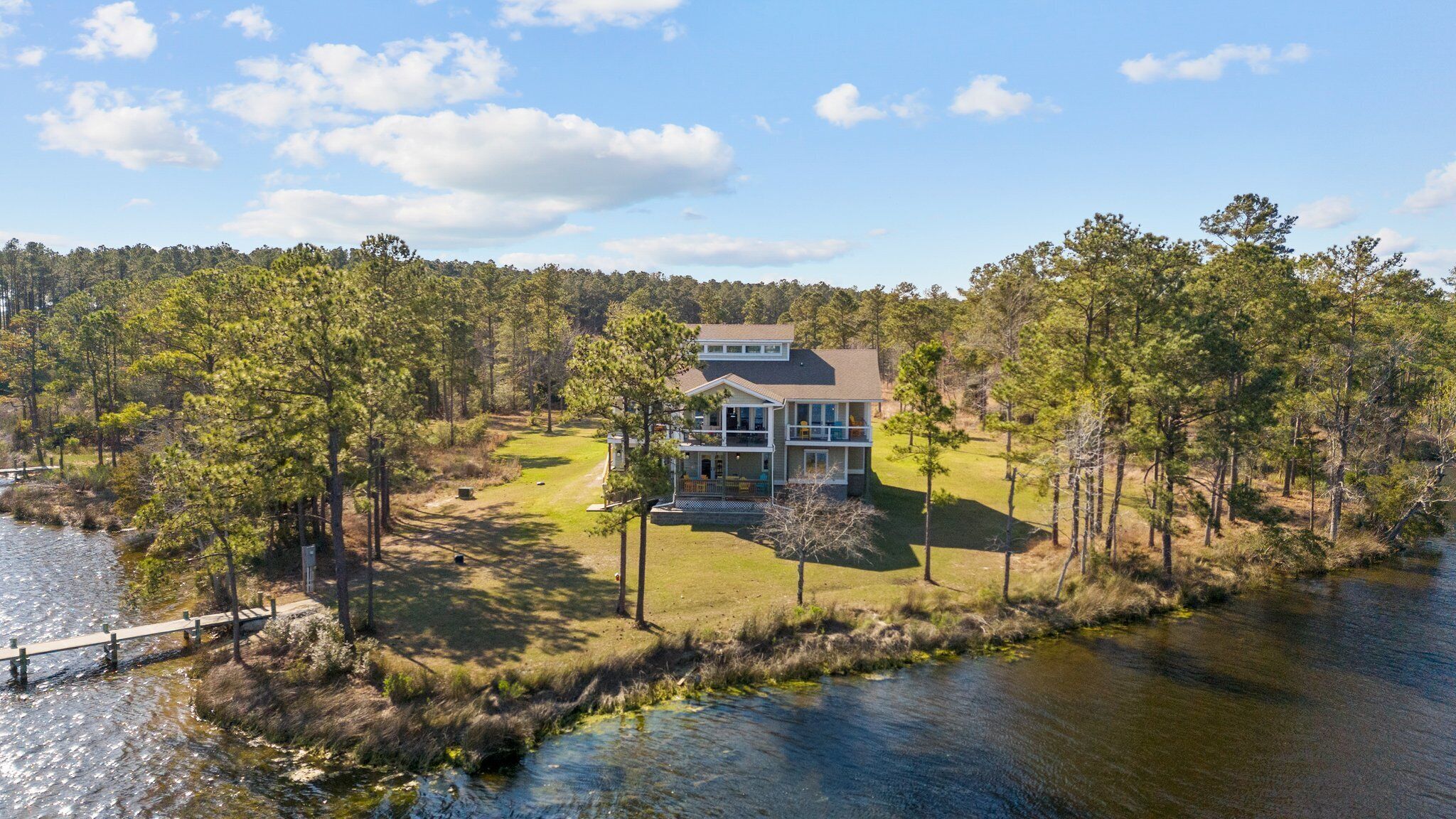 Une maison à deux étages dotée d’une grande terrasse, entourée d’arbres et bordée par un lac.