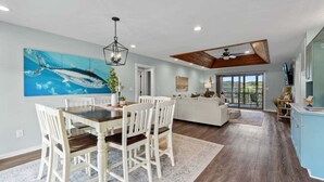 A spacious living area with a dining table and chairs, a large painting of a fish on the wall, and a view through a glass door to the outdoors. - Salty Bunker | 5200 ft to the beach | Private Pool (Kitty Hawk)