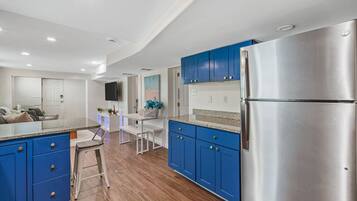 A modern kitchen with blue cabinets, a stainless steel refrigerator, and a dining area with a white table and bench.
