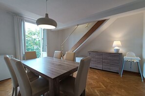 Un espace repas comprenant une table et des chaises en bois, une lampe, un plafond haut et une vue sur l’extérieur.