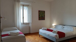 Habitació d'hotel amb dos llits, una finestra amb cortines blanques, un radiador i un quadre abstracte i colorit a la paret.
