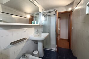Une salle de bain moderne avec un lavabo blanc, une cabine de douche en verre et une porte en bois donnant sur une autre pièce.